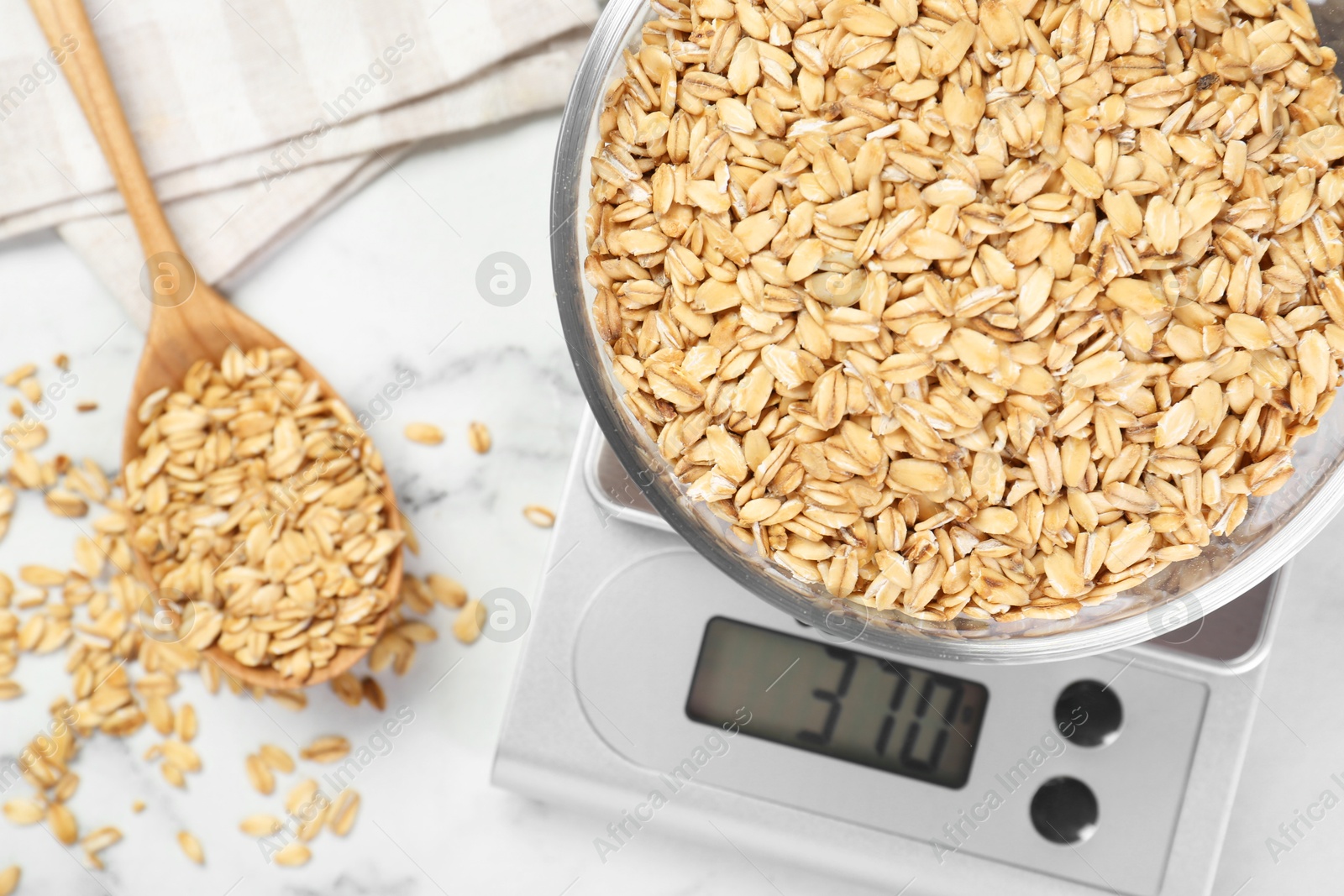 Photo of Kitchen scale with bowl of dry oats and spoon on white marble table, flat lay