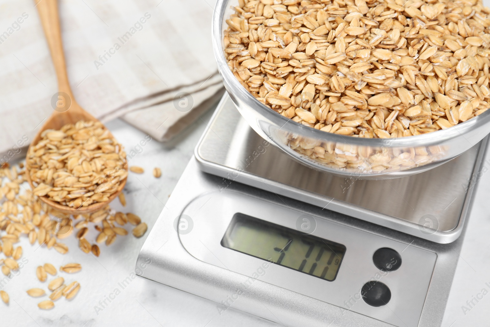 Photo of Kitchen scale with bowl of dry oats and spoon on white table, closeup