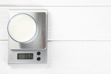 Kitchen scale with glass of milk on white wooden table, top view. Space for text