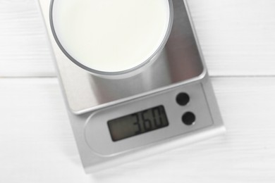 Kitchen scale with glass of milk on white wooden table, top view