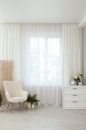 Soft armchair, chest of drawers, houseplant and window with curtains in room. Interior design