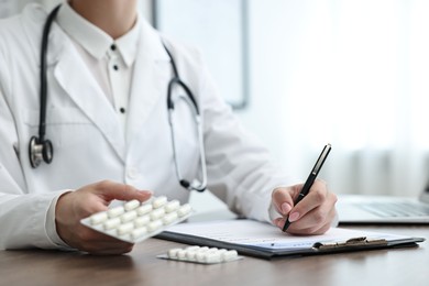 Photo of Doctor with pills writing prescription at wooden table in clinic, closeup