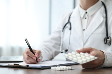 Photo of Doctor with pills writing prescription at wooden table in clinic, closeup