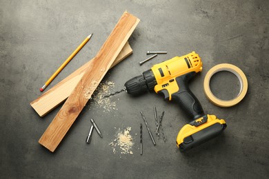 Photo of Modern cordless electric drill, sawdust, planks and bits on grey table, flat lay