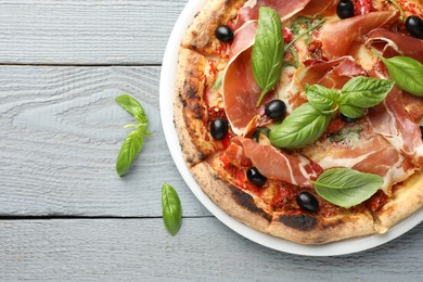 Photo of Tasty pizza with cured ham, olives, sun-dried tomato and basil on gray wooden table, top view. Space for text