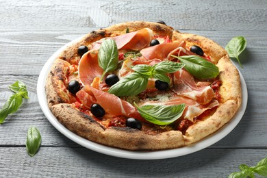 Photo of Tasty pizza with cured ham, olives, sun-dried tomato and basil on gray wooden table, closeup