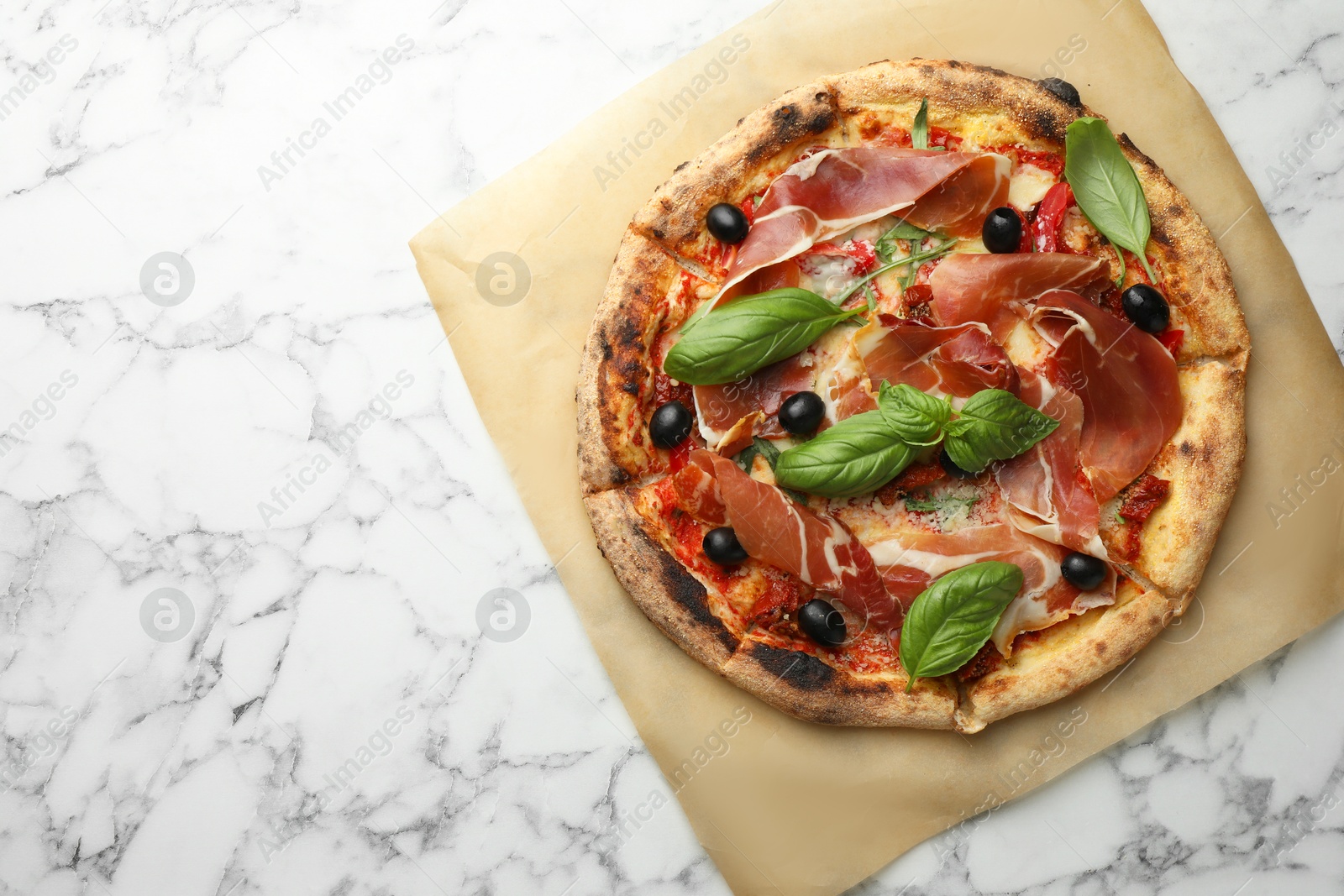 Photo of Tasty pizza with cured ham, olives, sun-dried tomato and basil on white marble table, top view. Space for text