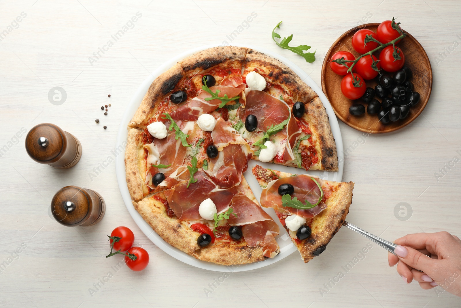 Photo of Woman taking piece of tasty pizza with cured ham, olives, mozzarella cheese and arugula at light wooden table, top view