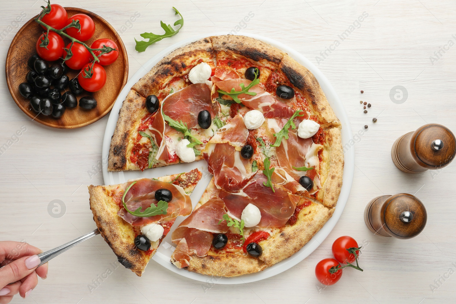 Photo of Woman taking piece of tasty pizza with cured ham, olives, mozzarella cheese and arugula at light wooden table, top view