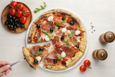 Photo of Woman taking piece of tasty pizza with cured ham, olives, mozzarella cheese and arugula at light wooden table, top view