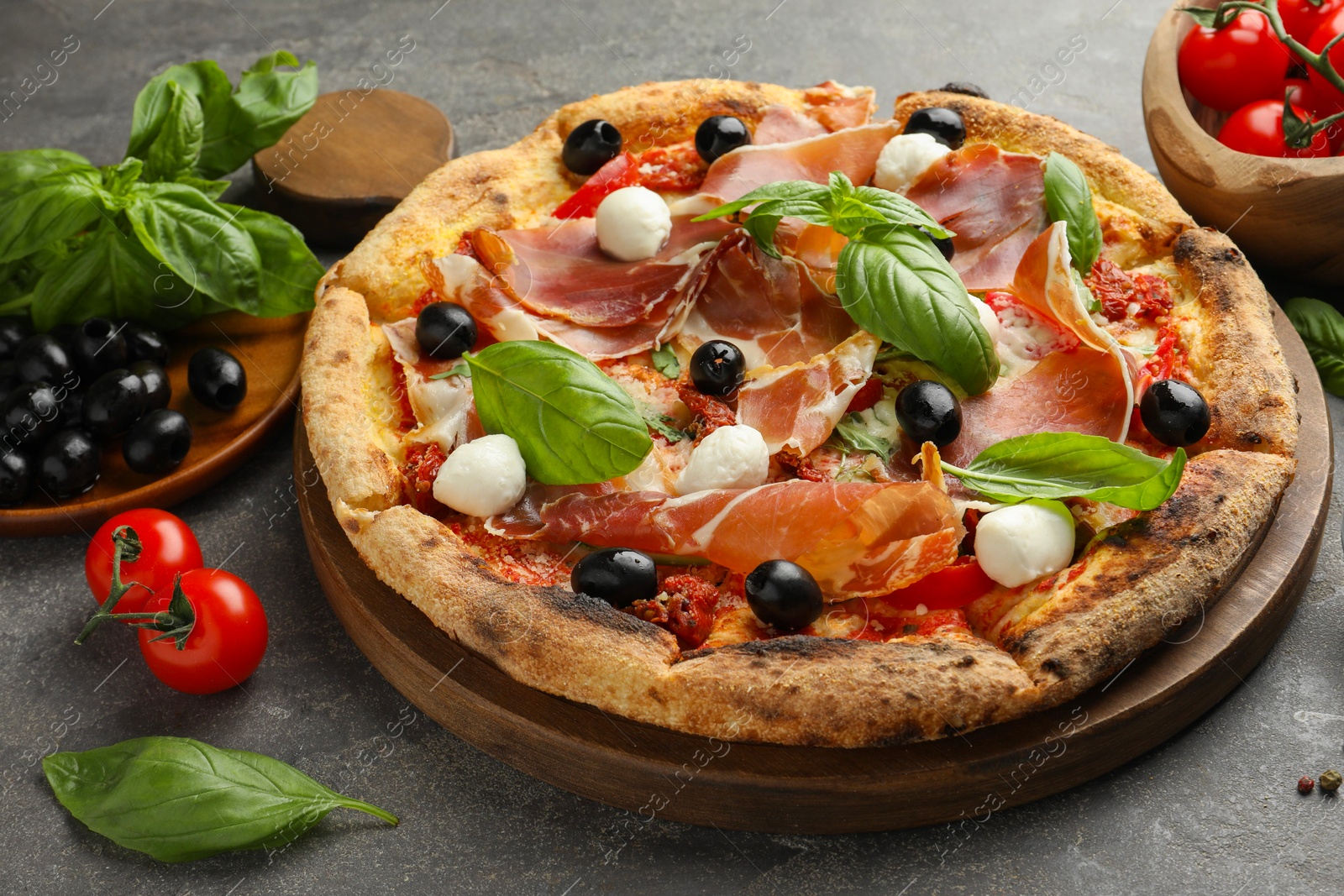 Photo of Tasty pizza with cured ham, olives, mozzarella cheese, tomatoes and arugula on gray table