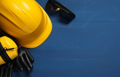 Photo of Hard hat, earmuffs, protective gloves and goggles on blue wooden background, flat lay. Space for text
