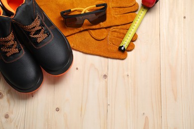 Photo of Pair of working boots, protective gloves, goggles and tape measure on wooden background, flat lay. Space for text