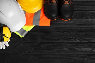 Photo of Pair of working boots, hard hats, protective gloves, earmuffs and reflective vests on black wooden background, flat lay. Space for text