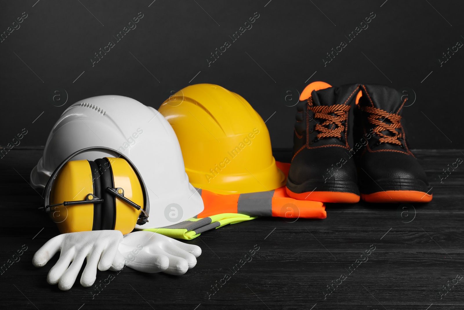 Photo of Pair of working boots, hard hats, protective gloves, earmuffs and reflective vests on black wooden surface against gray background