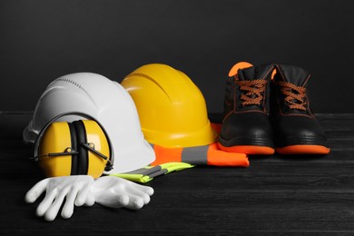 Photo of Pair of working boots, hard hats, protective gloves, earmuffs and reflective vests on black wooden surface against gray background