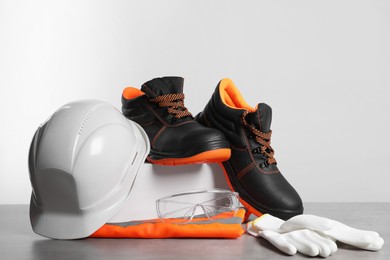 Photo of Hard hat, pair of working boots, protective gloves, goggles and reflective vest on gray surface against light background