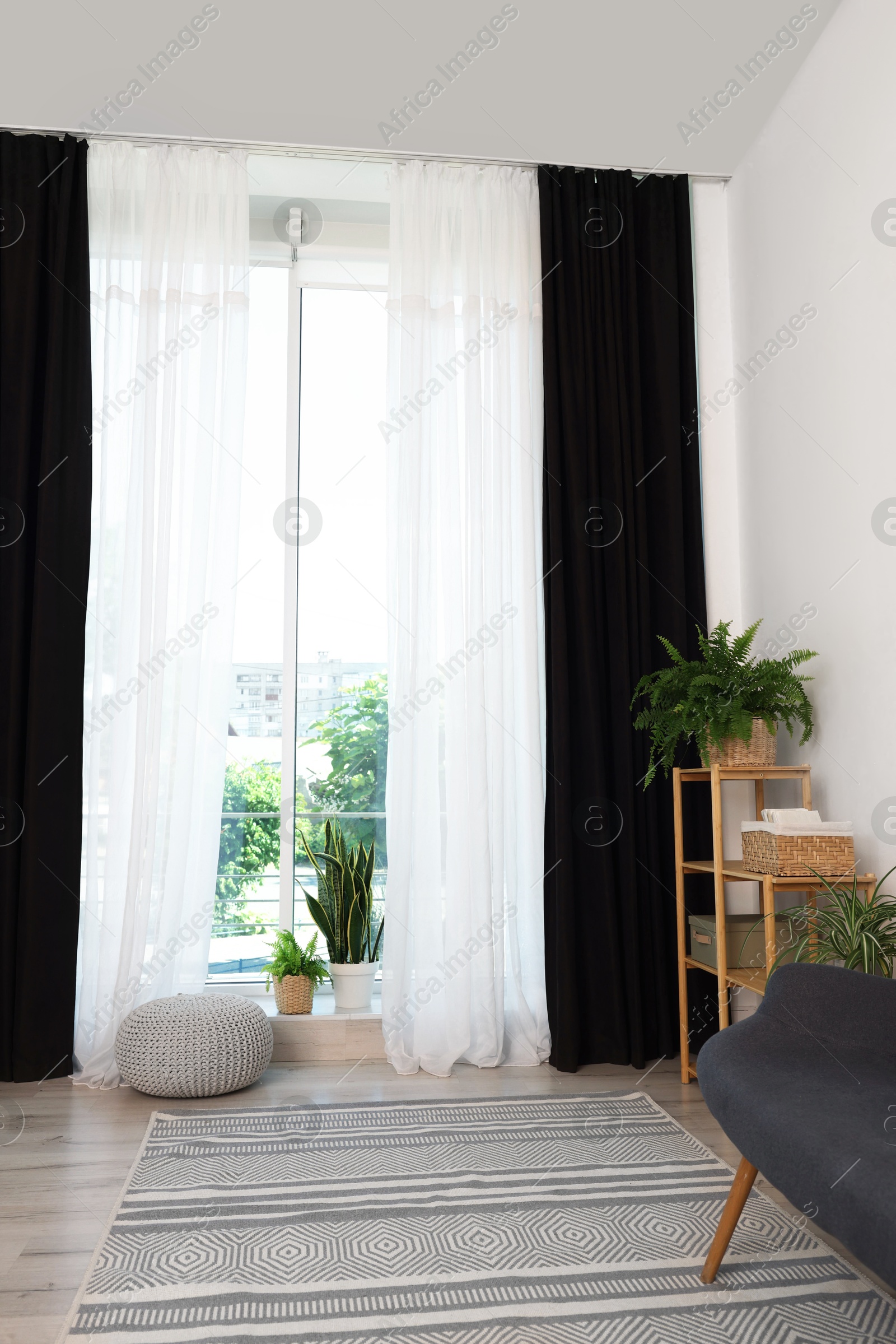 Photo of Shelving unit, sofa, green houseplants, pouffe and elegant curtains in room. Interior design