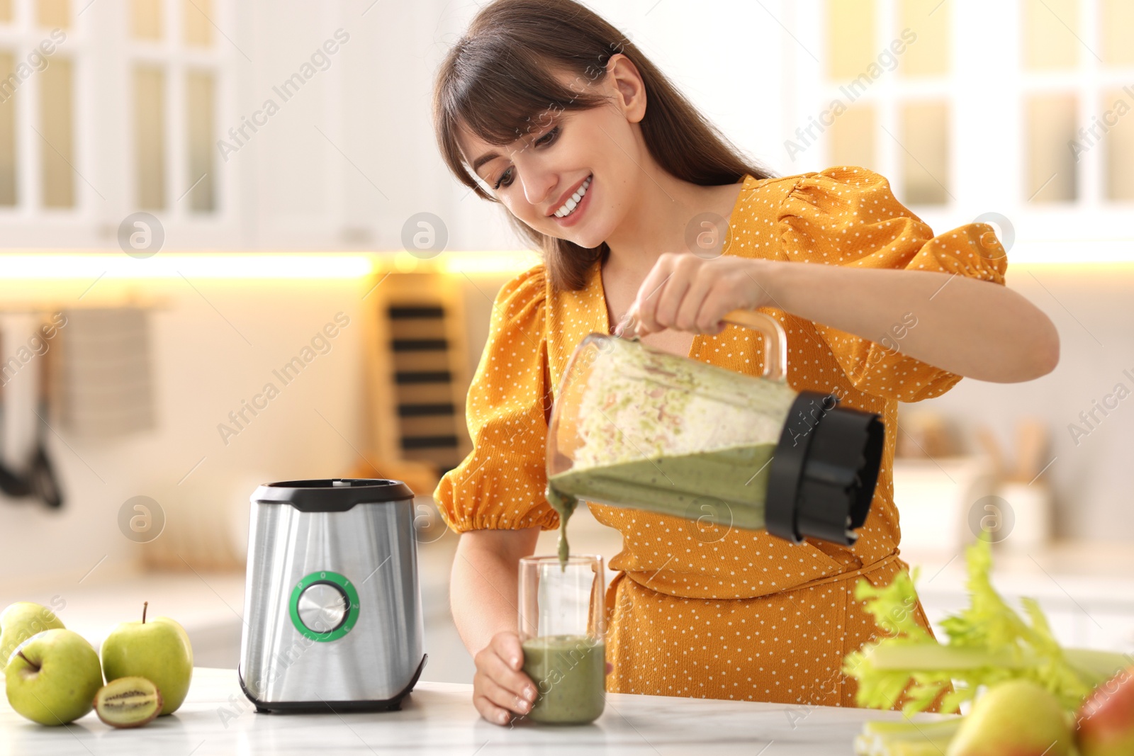 Photo of Young woman pouring fresh smoothie from blender cup into glass at white marble table in kitchen