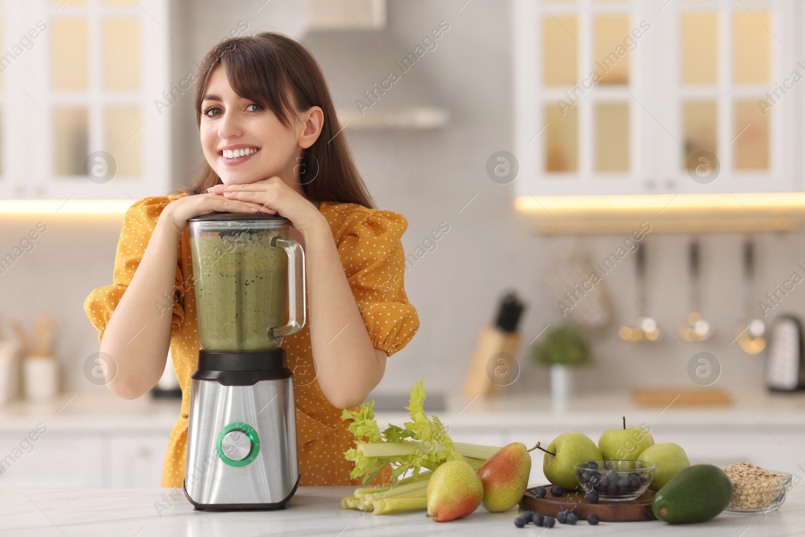 Photo of Young woman making delicious smoothie with blender at white marble table in kitchen. Space for text