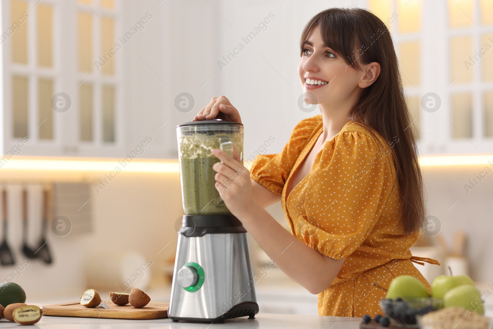 Photo of Young woman making delicious smoothie with blender at white marble table in kitchen