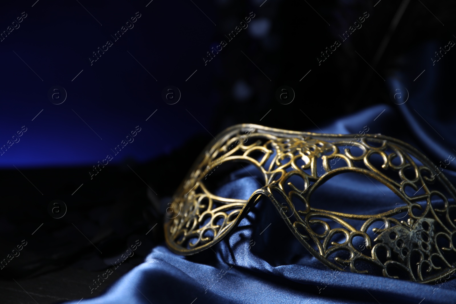 Photo of Beautiful carnival mask on blue fabric in darkness, closeup