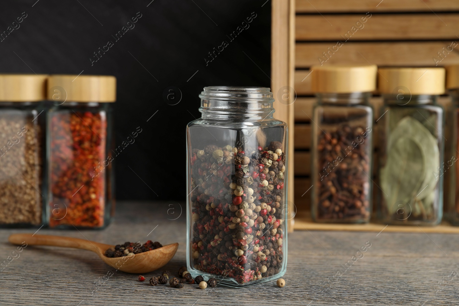 Photo of Peppercorns in glass jar, spoon and other spices on grey wooden table, closeup