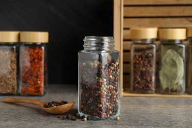 Peppercorns in glass jar, spoon and other spices on grey wooden table, closeup