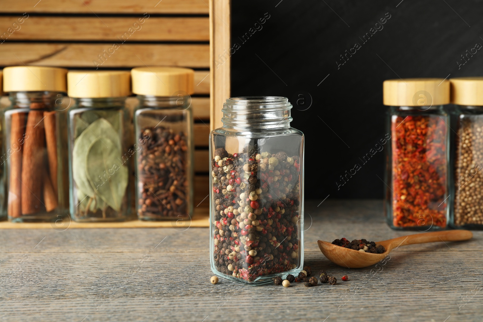 Photo of Peppercorns in glass jar, spoon and other spices on grey wooden table, closeup