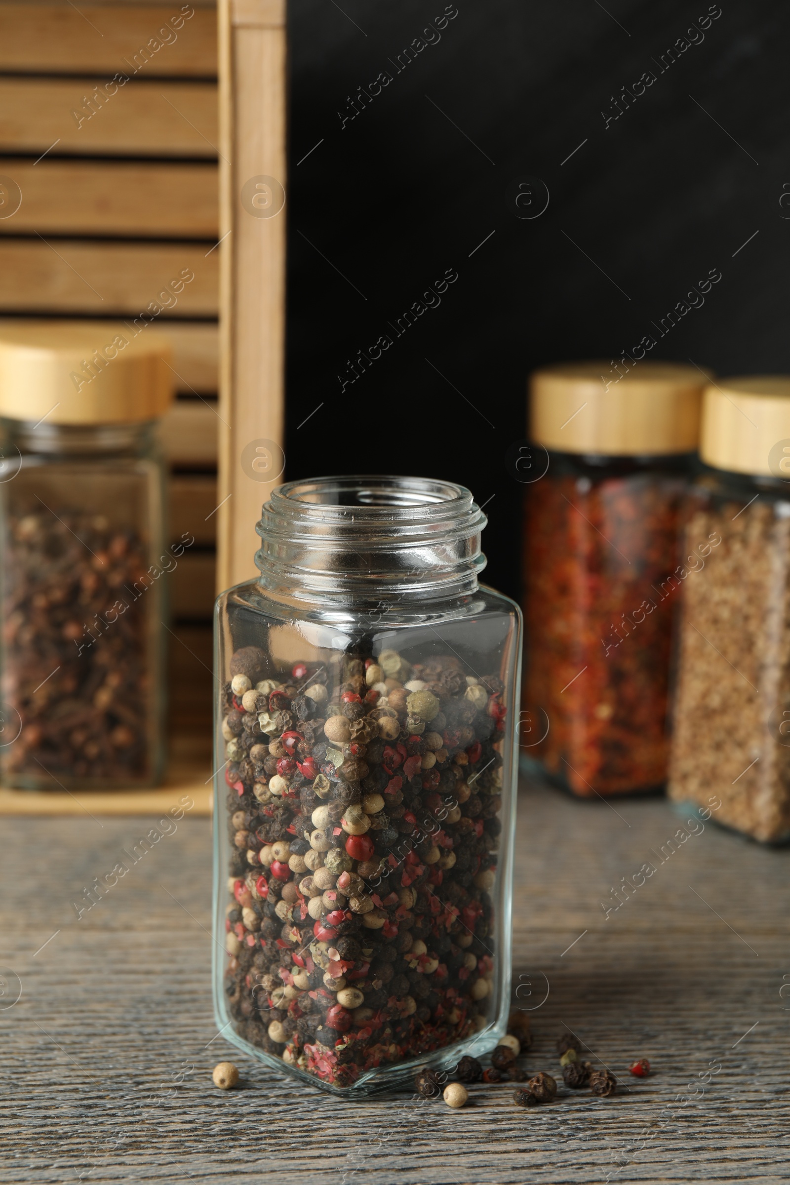 Photo of Peppercorns in glass jar and other spices on grey wooden table, closeup