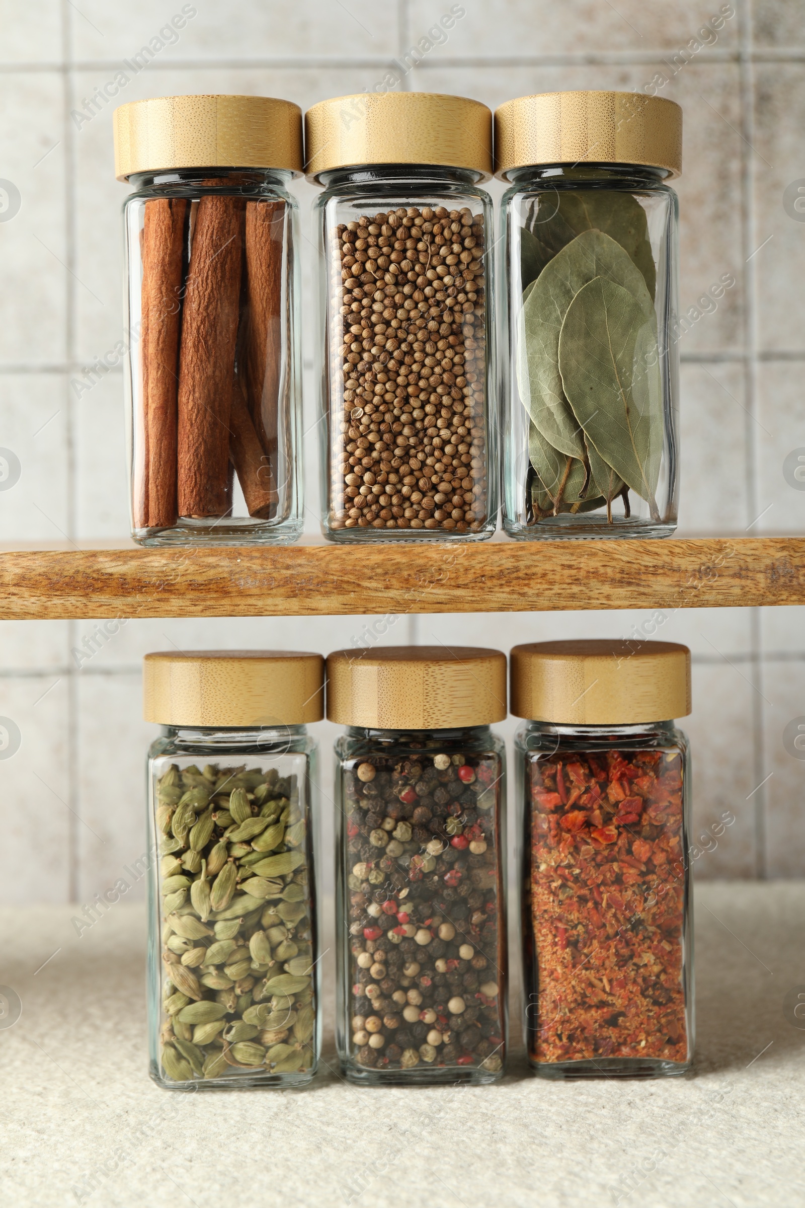 Photo of Different spices in glass jars on light textured table under shelf with ones, closeup