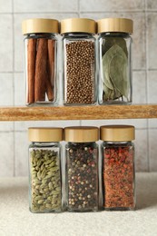 Photo of Different spices in glass jars on light textured table under shelf with ones, closeup