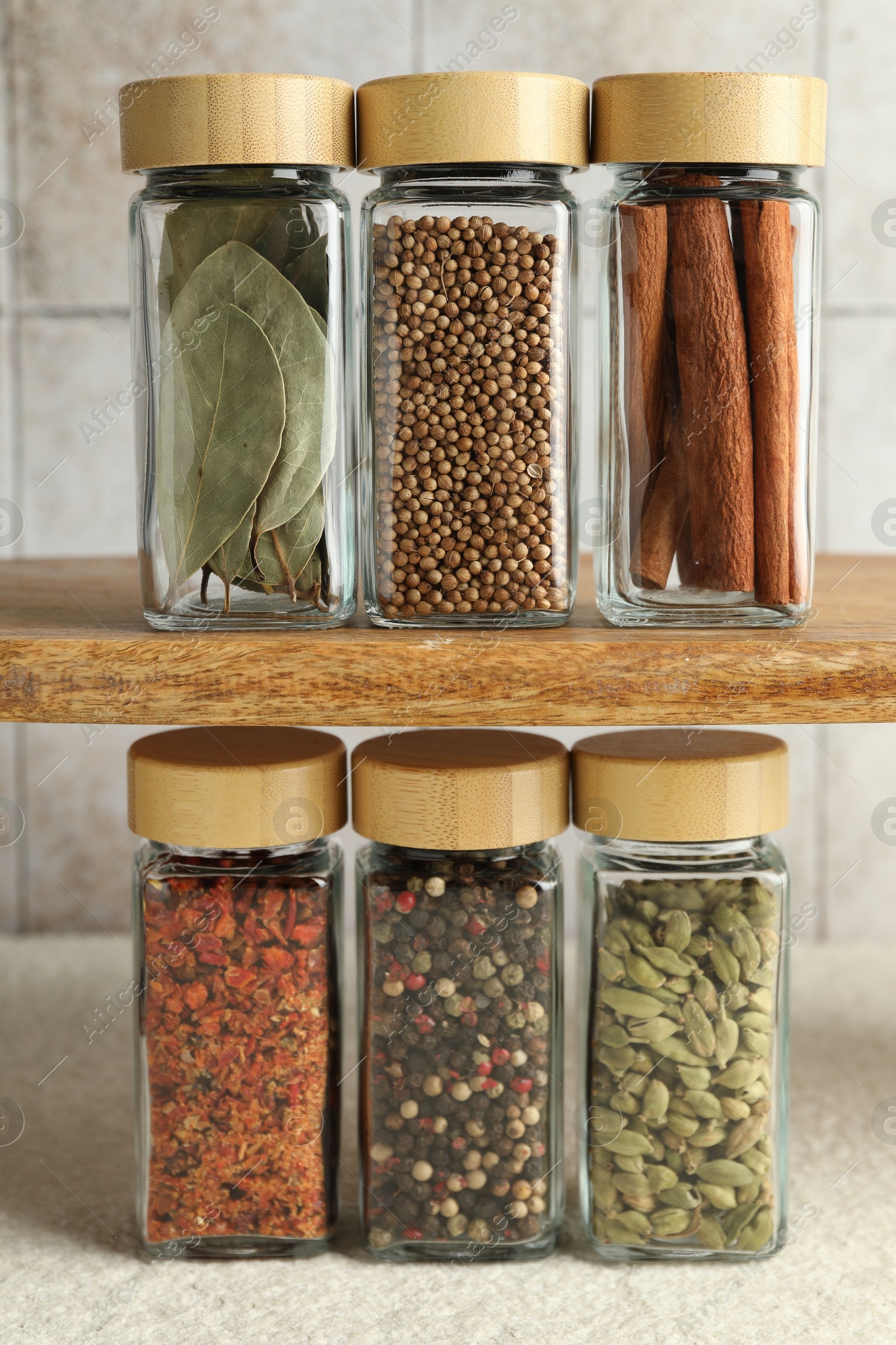 Photo of Different spices in glass jars on light textured table under shelf with ones, closeup