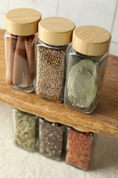 Photo of Different spices in glass jars on light textured table under shelf with ones, closeup