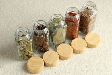 Photo of Different spices in glass jars and lids on light textured table, closeup