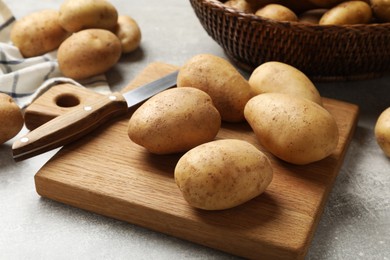 Photo of Many fresh potatoes, board and knife on grey table, closeup