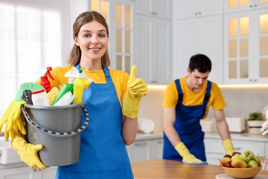 Photo of Cleaning service worker holding bucket with supplies and showing thumbs up in kitchen