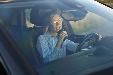 Photo of Smiling woman in glasses with smartphone driving car, view through windshield