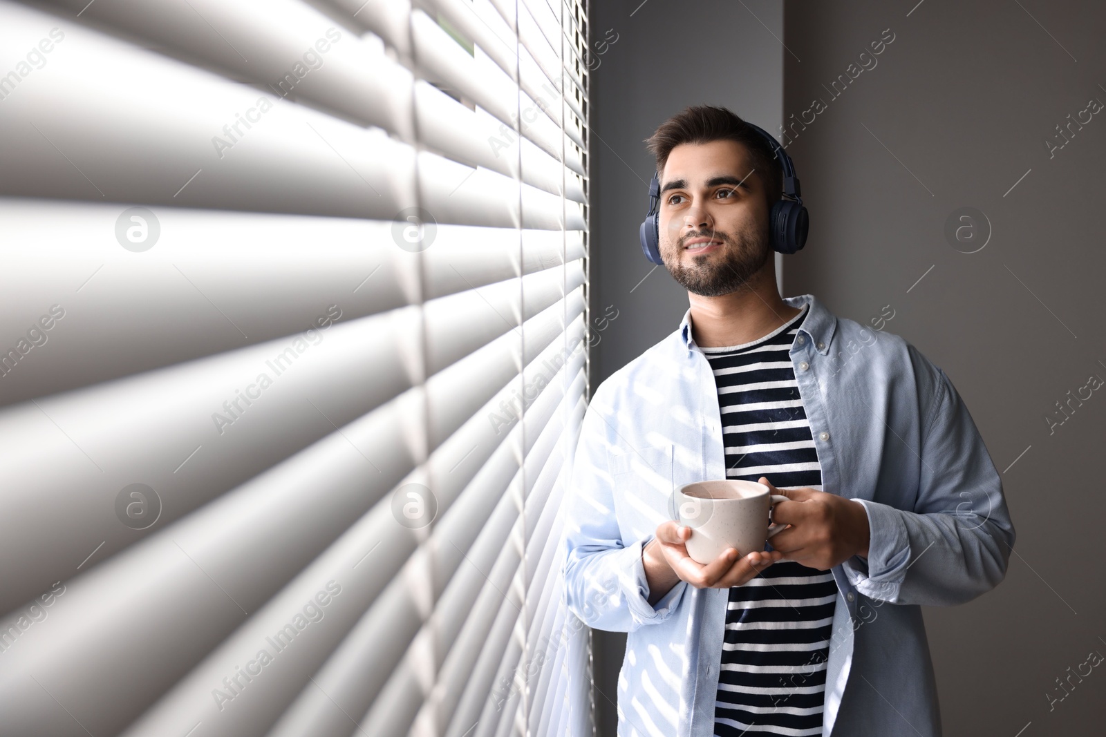 Photo of Man with headphones and cup of drink listening to music near window blinds at home, space for text