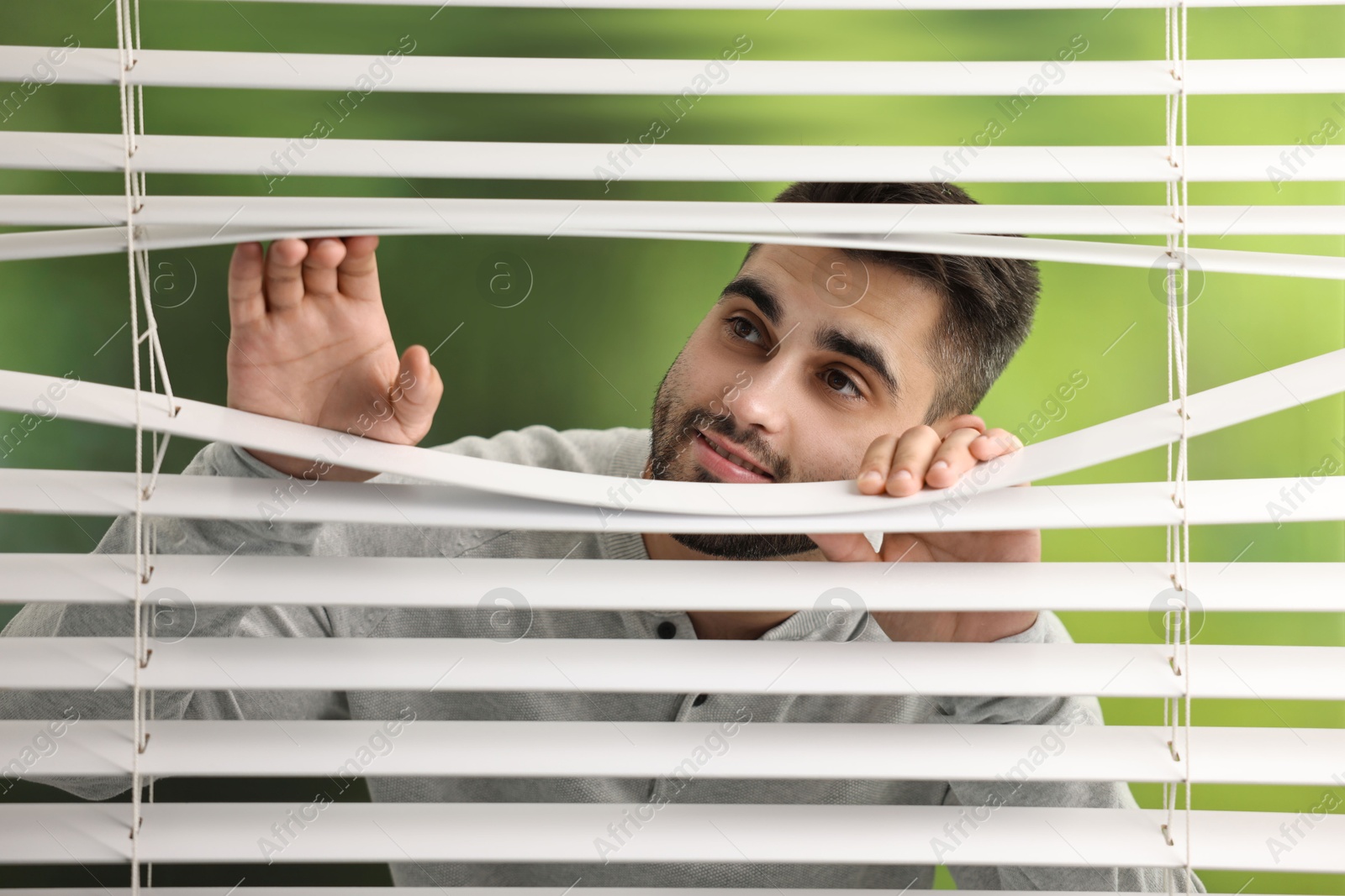 Photo of Young man looking through window blinds on blurred background