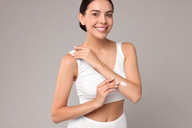 Smiling woman applying cream onto hand on grey background. Body care