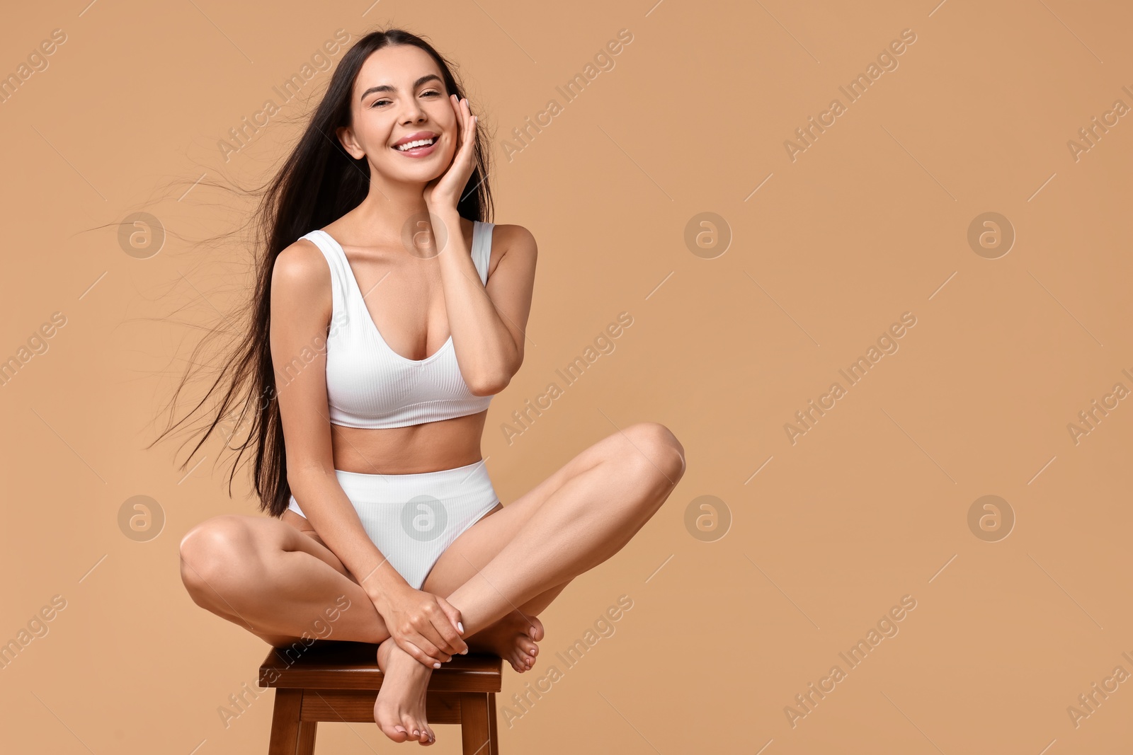 Photo of Smiling woman with perfect skin in underwear posing on stool against beige background, space for text. Body care