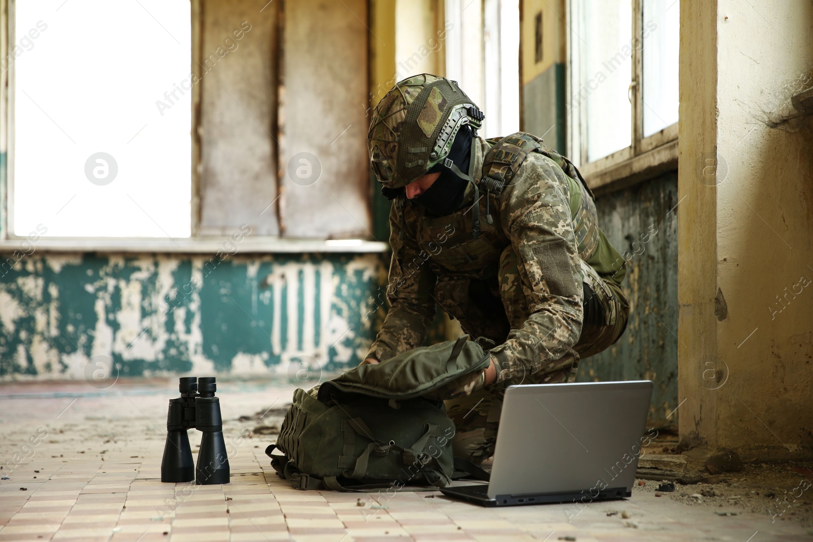 Photo of Military mission. Soldier in uniform using laptop and binoculars inside abandoned building