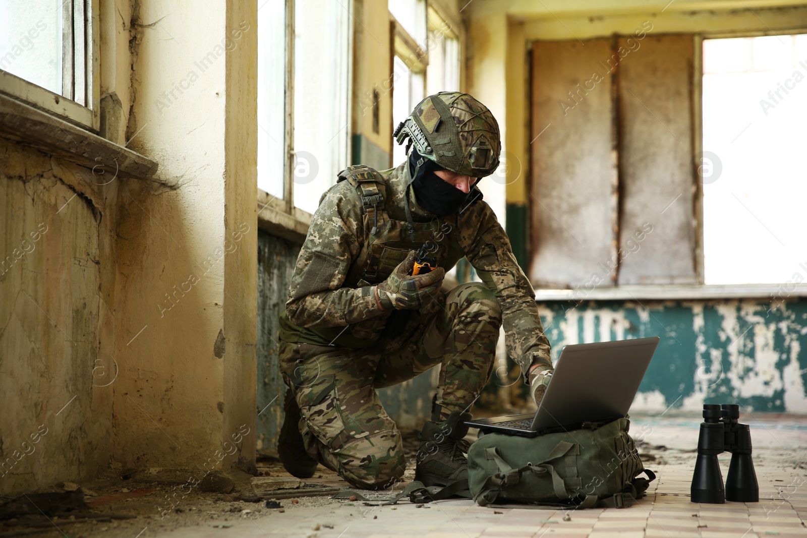 Photo of Military mission. Soldier in uniform using laptop and binoculars inside abandoned building