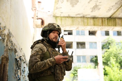 Photo of Military mission. Soldier in uniform with drone controller and radio transmitter near abandoned building outdoors, low angle view. Space for text