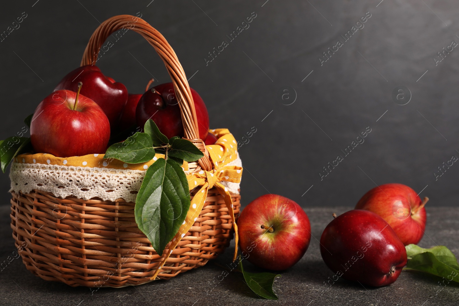 Photo of Ripe red apples and leaves in wicker basket on dark grey table. Space for text