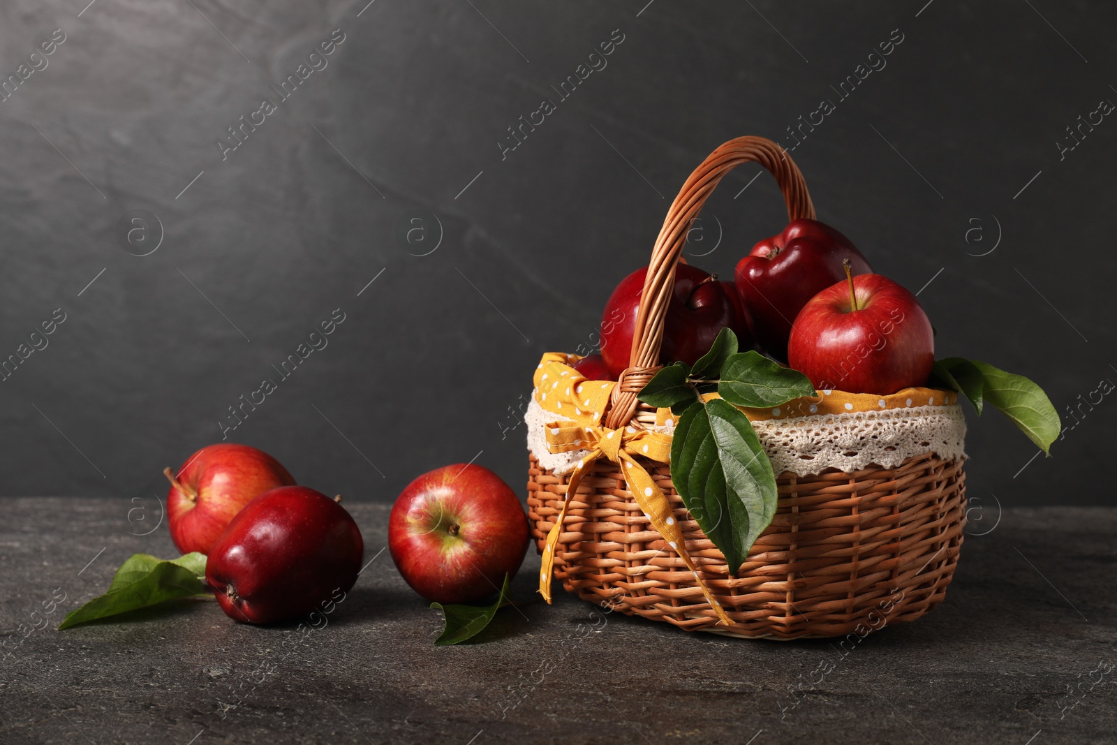 Photo of Ripe red apples and leaves in wicker basket on dark grey table. Space for text