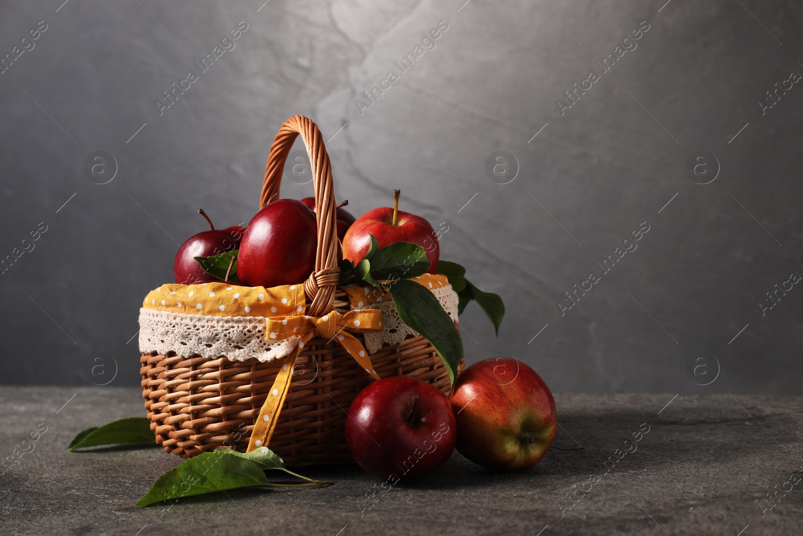 Photo of Ripe red apples and leaves in wicker basket on dark grey table. Space for text