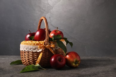 Photo of Ripe red apples and leaves in wicker basket on dark grey table. Space for text