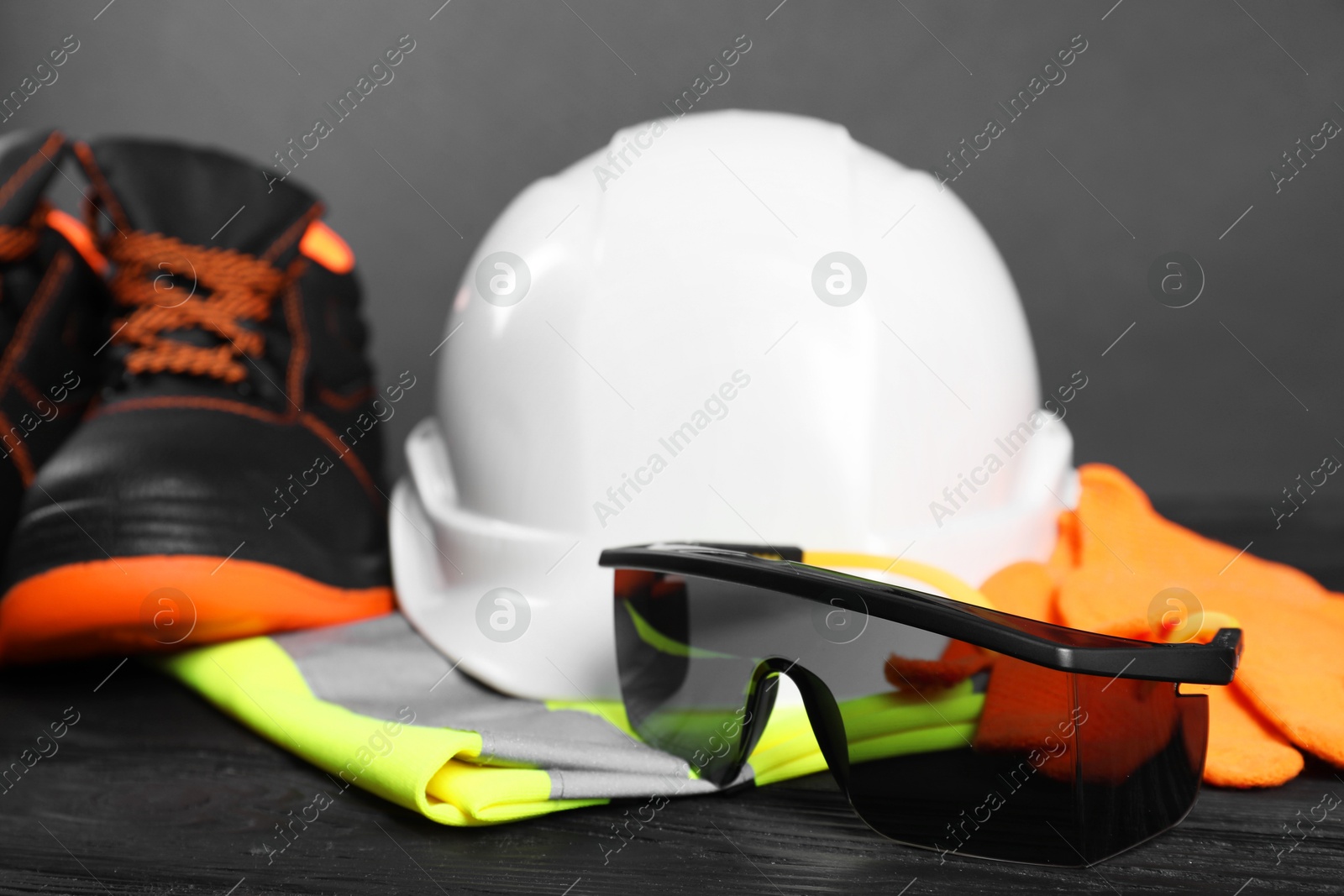 Photo of Pair of working boots, hard hat, protective gloves, reflective vest and goggles on black wooden background, closeup